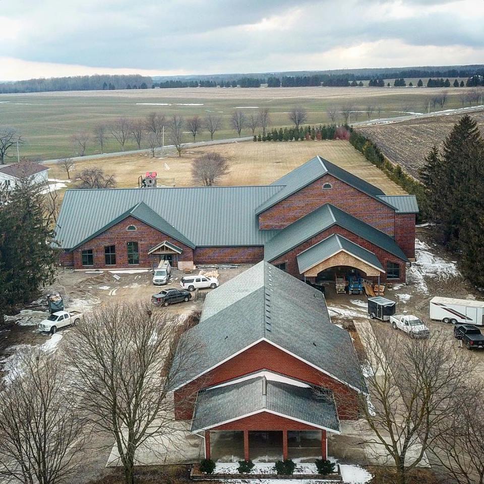 Aerial view of a new church building.  The new church as a steel roof, carport, steel framing for the sanctuary.  The old church is shown in front of the new church.  Mapleview Mennonite Church located in the countryside.