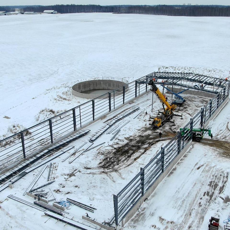 Pre-engineered shed is being installed on a farm.  A crane is being used to install the steel roof.  Aerial view from a drone.  Installation is during winter and snow is on ground.  Both a crane and manlift are being used to install the pre-eng building.
